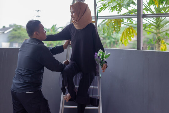 Asian Muslim couple playfully interacting around a stepladder with flowers on a balcony.