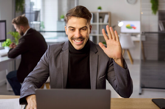 Businessman online video call chat on laptop. Smiling professional greets clients with a wave in a modern office. Clear concise communication matters. Virtual business connection for teamwork.