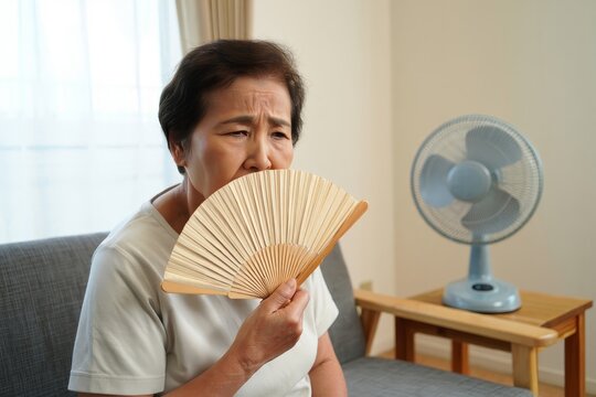 Elderly asian woman fanning herself with a traditional hand fan due to hot weather indoors