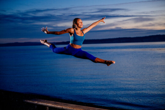 Hispanic woman performing yoga acrobatic leap by the ocean at sunset