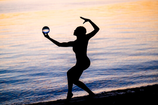 Hispanic woman practicing yoga acrobatics with ball on shore at sunset