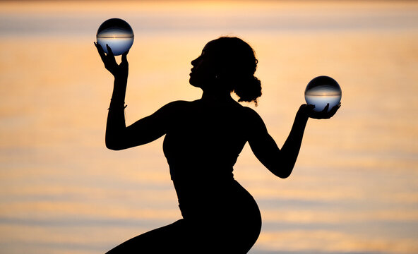 Hispanic woman practicing yoga holding glass balls at sunset by water