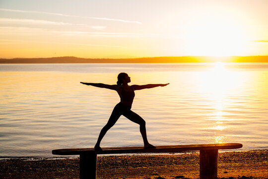 Hispanic woman practicing yoga warrior pose on bench at beach sunset