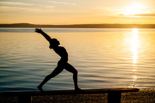 Hispanic woman performing yoga stretch pose by lake at sunset in USA