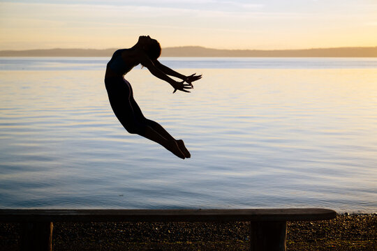 Hispanic woman jumping acrobatically over bench by lake at sunset USA