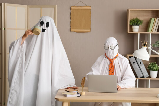 Halloween office party and spooky work environment, people in ghost costumes working on laptop at desk