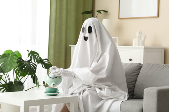 Person in a ghost costume pouring tea into a cup on a white table in a living room, the ghost wears white gloves while sitting on a gray sofa in a brightly lit living room with a large green monstera