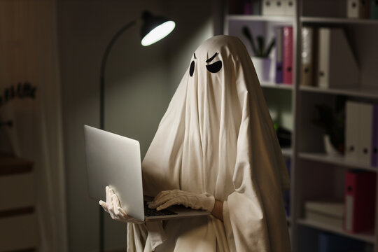 Ghost in white sheet costume with angry face using laptop in dark office at night, internet security and privacy, this image internet security online privacy and cybersecurity concepts