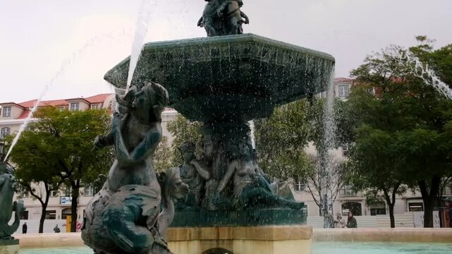 The ornate Rossio Square fountain stands in central Lisbon, Portugal, surrounded by historic buildings and patterned pavement under a cloudy sky