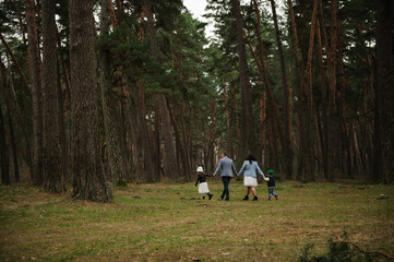 Happy family with mom, dad, son, and daughter walking together through a vast pine forest. Back view. © Serhii