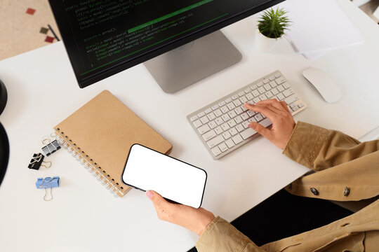 Female programmer working with mobile phone and computer on table in office, top view
