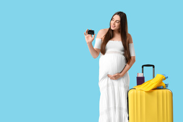 Young pregnant woman holding credit card near suitcase and flippers on blue background. Travel concept © Pixel-Shot