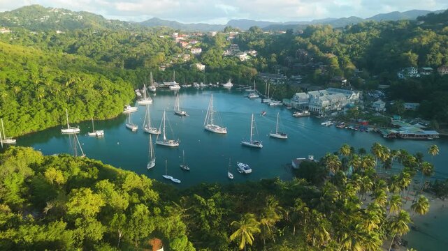 Aerial drone parallax shot reveals sailboats anchored in sheltered Marigot Bay harbor with palm trees and hillside village in St Lucia