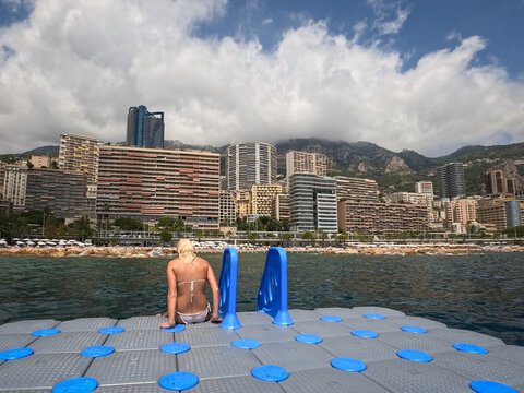 Young blonde woman in a white bikini sitting on a modular floating platform pontoon with the urban Monaco cityscape and mountains in the background.