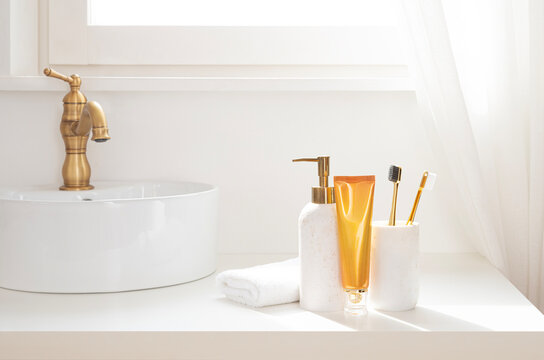 Bathroom counter top interior with white vessel sink and gold faucet, amber color cream tube, soap dispenser, folded fluffy towel and toothbrush holder in soft daylight on window curtain background