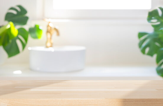 Empty wooden table in dappled sunlight on blurred background of white vessel sink with gold faucet and green plant tropical leaves