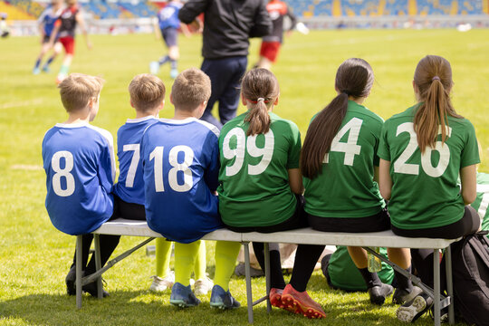 Junior Soccer Substitutes in Blue and Green Numbered Shirts Sitting on Sideline Bench - Youth Football Team Support and Substitution Strategy