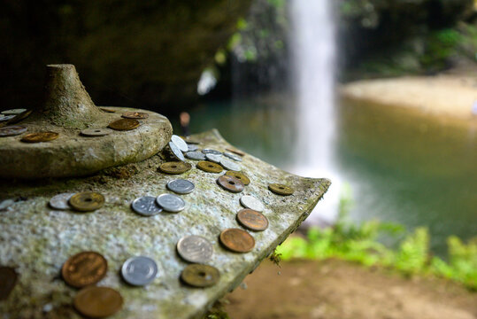 Close-up macro view of Japanese coins offered as donations on a weathered stone jizo shrine with a waterfall in the background