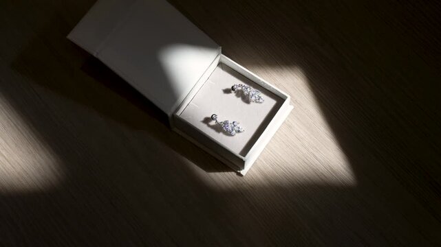 A close look at earrings in a box. Sunlight shines on the box and illuminates the earrings. The setting is a simple wooden table.