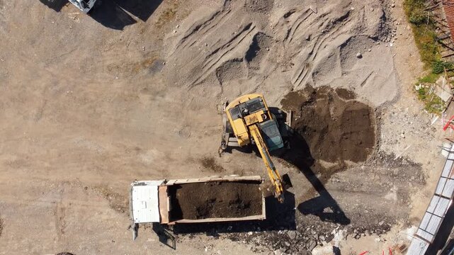 Aerial top-down view of a yellow excavator scooping up earth and loading it into the back of a large white dump truck at a busy construction site, representing industrial groundwork