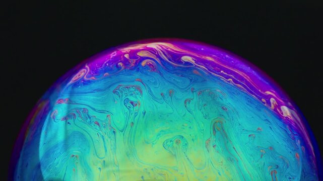 The movement of fluid within the bubble's film creates dynamic abstract patterns, swirls, and a colorful array of hues&mdash;such as blue, green, purple, and yellow&mdash;set against a deep black background.