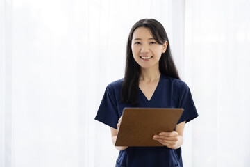 Obraz premium Young Japanese female doctor in scrubs holding a clipboard and smiling at the camera by a bright window