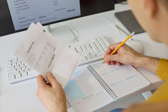 Man reviews electricity bill and budget on computer. He records expenses in a notebook while managing home finances, planning payments and tracking utility costs. Focus on smart money management.