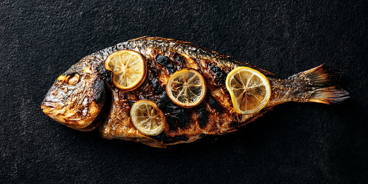 A whole grilled fish topped with lemon slices on a dark textured surface. The fish skin is charred and crispy, indicating a delicious and flavorful meal