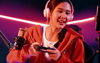 Host channel of smiling beautiful Asian girl streamer with joystick playing online game wearing headphones paste talking with viewers media online. Esport skilled team players in neon room. Stratagem. © InfiniteFlow