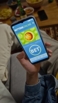 Vertical shot of young adult man tapping sports betting application on smartphone screen beside friends at home. Clip suited for mobile gambling marketing