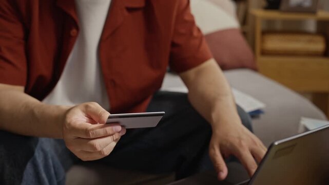 Asian young adult man holding credit card and typing on laptop to pay utility bills at home. Footage is perfect for personal finance, online banking, and debt management ads