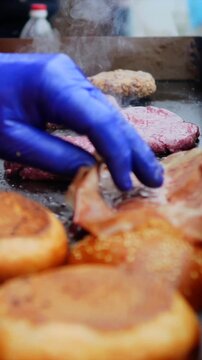 Grill master turns meat patties beside rows of burger buns and cheese slices. Blue gloves move over sizzling surface while smoke and steam drift through outdoor cooking, seasoning patties on griddle.