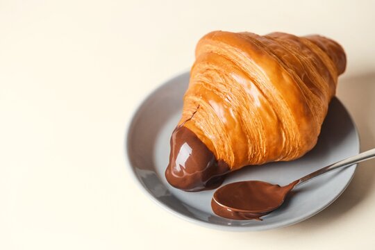 Fresh croissant dipped in a chocolate cream with a spoon on a gray plate, beige background. Copy space, Side view