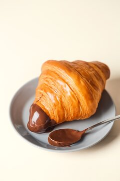 Fresh croissant dipped in a chocolate cream with a spoon on a gray plate, beige background. Side view