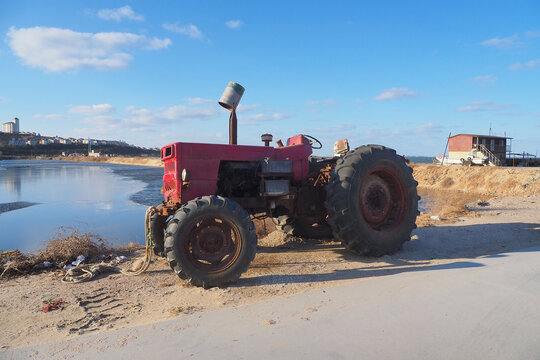 Old red tractor on sandy shore beside calm water under blue sky