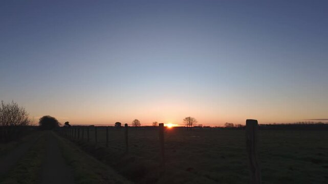 Tranquil sunrise over a peaceful rural landscape with a fence line and path. Cinematic footage of a new day dawning in the countryside.