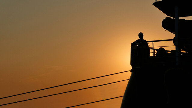 silhouette of a line handler man standing on stern working on a ship casting off rope from dock against sunset sky with space for text