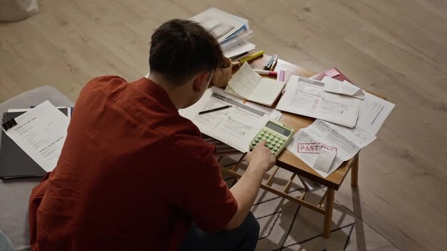 Asian young adult man calculating household expenses and managing past due utility bills at home, high angle shot. Perfect for financial planning services or debt relief advertisements