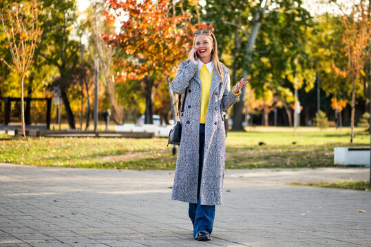 Beautiful and confident young businesswoman walking through a city park in autumn while talking on her smartphone and enjoying a busy professional day.