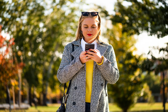 Beautiful and successful young businesswoman texting on her smartphone while walking through a city park during colorful autumn weather.