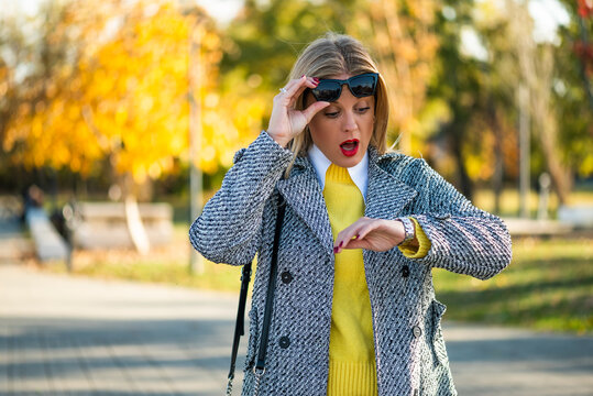 Worried businesswoman checking her wristwatch while walking through a city park in autumn, feeling stressed about a busy schedule and approaching deadline.
