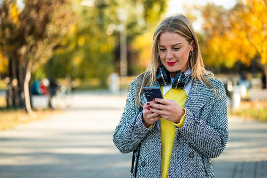 Beautiful and elegant businesswoman wearing wireless headphones and typing on her smartphone while standing in the city park during autumn day.