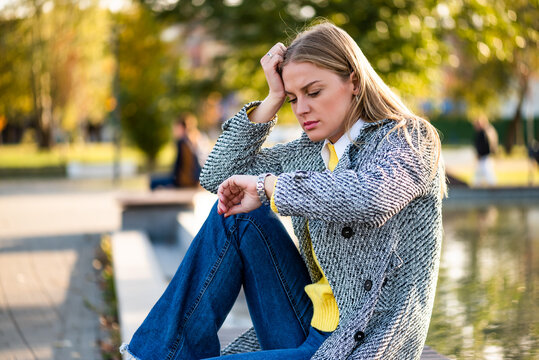 Stressed professional woman sitting in a public park, looking at her watch with frustration and anxiety while waiting and worrying about time.