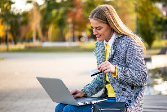Freelance businesswoman using laptop in the city park while managing online purchases and financial activities on a sunny autumn day.