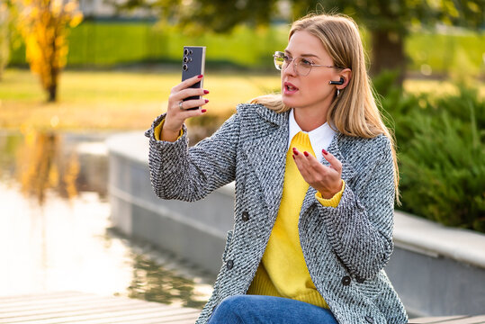 Stylish businesswoman with eyeglasses working remotely outdoor, using smartphone and headphones to stay connected during an online meeting on a autumn day.