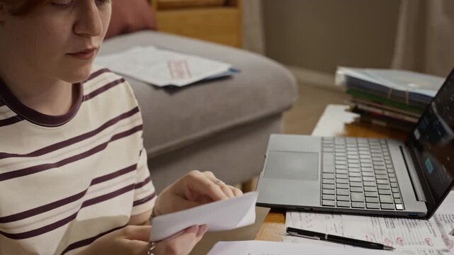 Caucasian young adult woman opening envelope and reading utility bill next to laptop. Footage is perfect for financial planning, banking services, and debt management advertisements