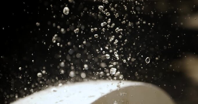 A close-up, 4K slow motion of water droplets spraying and freezing mid-air above a stone fountain basin against a soft, out-of-focus background of green foliage.