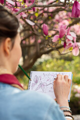 An artist draws a botanical sketch in a notebook beneath a blooming magnolia tree in Paris. The over-the-shoulder shot captures the drawing process in a vertical frame. © dsbedarieva