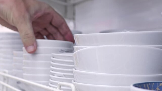 Man Selecting White Saucers for Purchase in Household Store. Close view of fingers choosing porcelain dishes from a shelf. Concept of consumer decision, kitchenware retail and everyday home use.