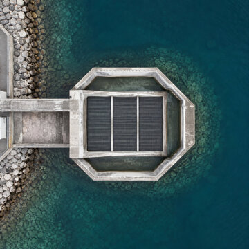 Aerial top down view of a concrete seawater intake structure with steel screens surrounded by turquoise ocean and rock armour at a coastal desalination site
